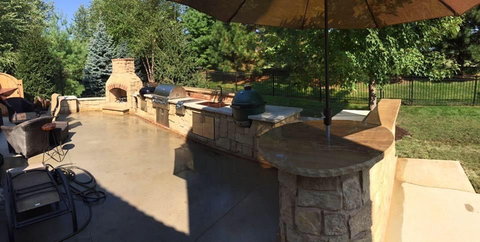 Backyard patio with stone fire pit, umbrella, and lounge chairs beside a lawn and trees.