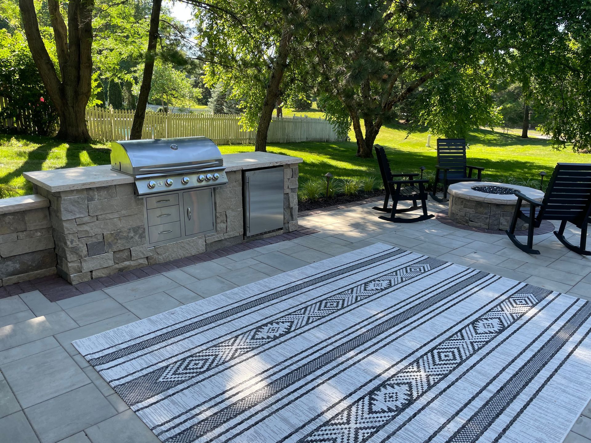 Stone outdoor grill and seating area with a striped rug on a patio under trees