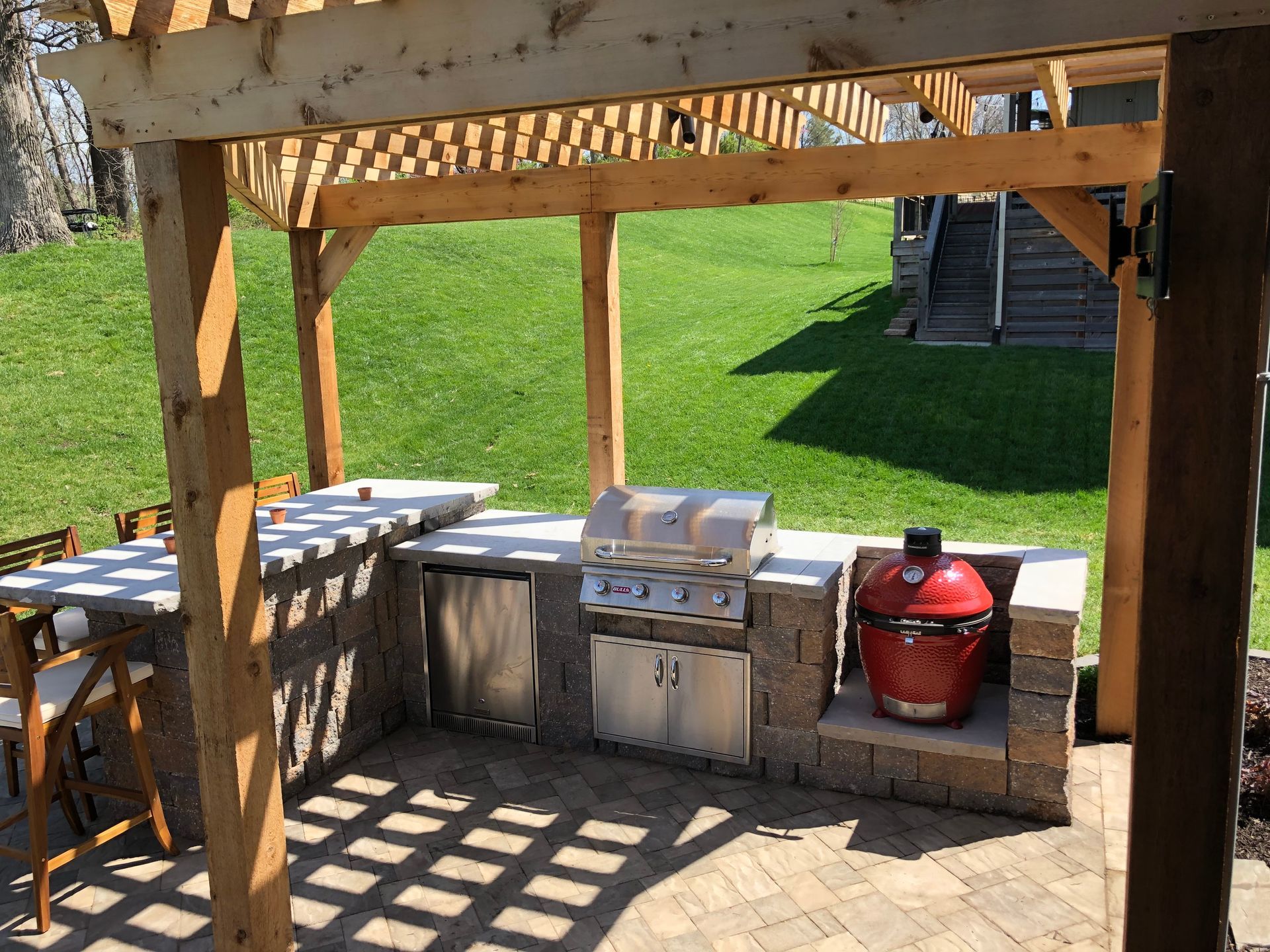 Outdoor patio kitchen under a wooden pergola with grill, sink, counter, and red smoker on a brick wall
