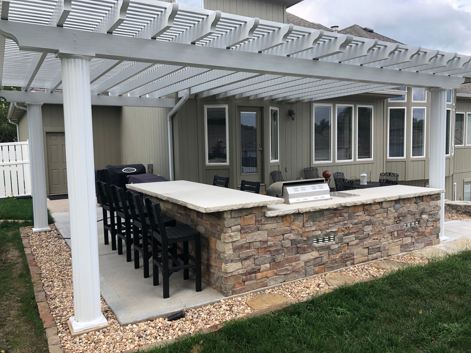 Patio with white pergola, stone bar, and black stools beside a house backyard