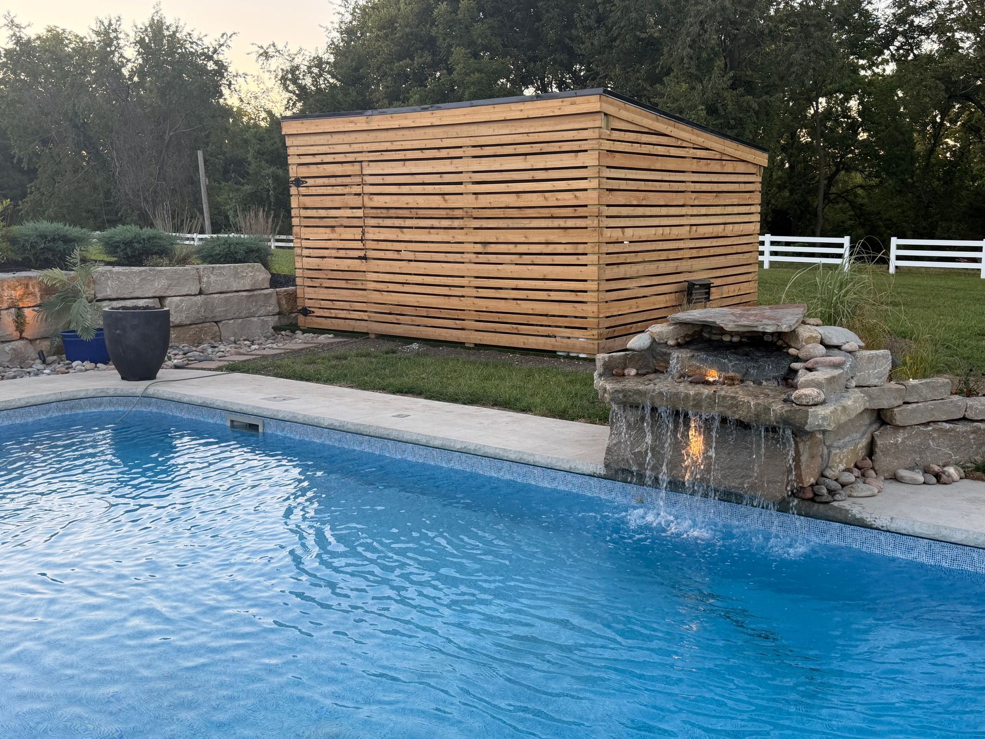 Backyard pool with stone waterfall, wooden privacy screen, and blue water at dusk