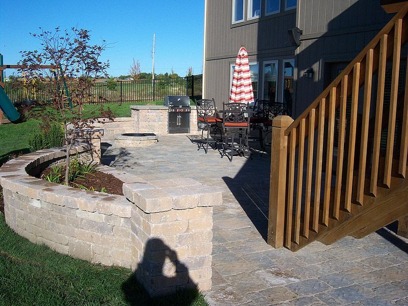 Patio with grill, seating, and retaining wall. Adjacent to house with wooden stairs and umbrella.