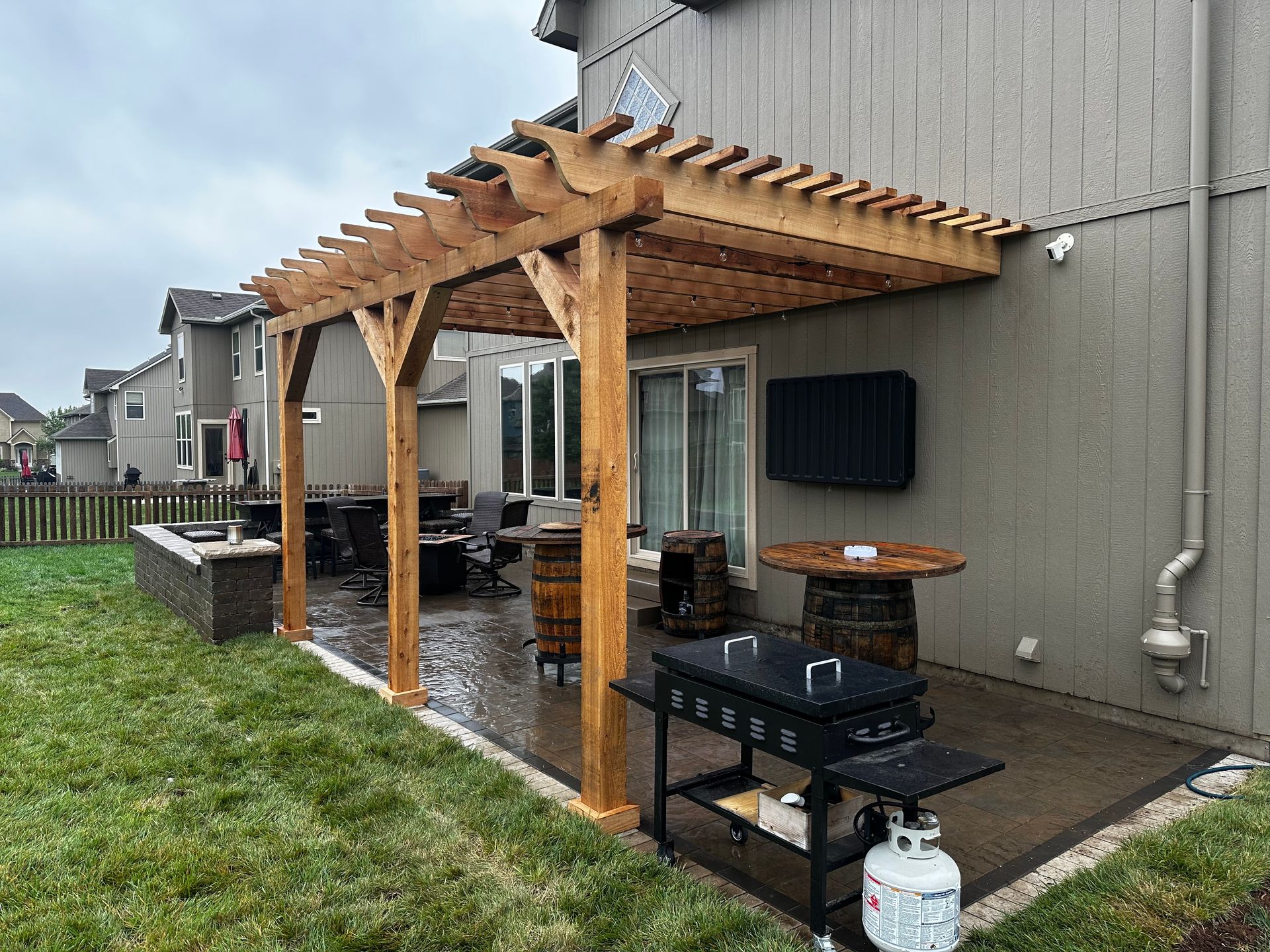 Wooden pergola over a wet backyard patio with grill, seating, and a propane tank beside a house
