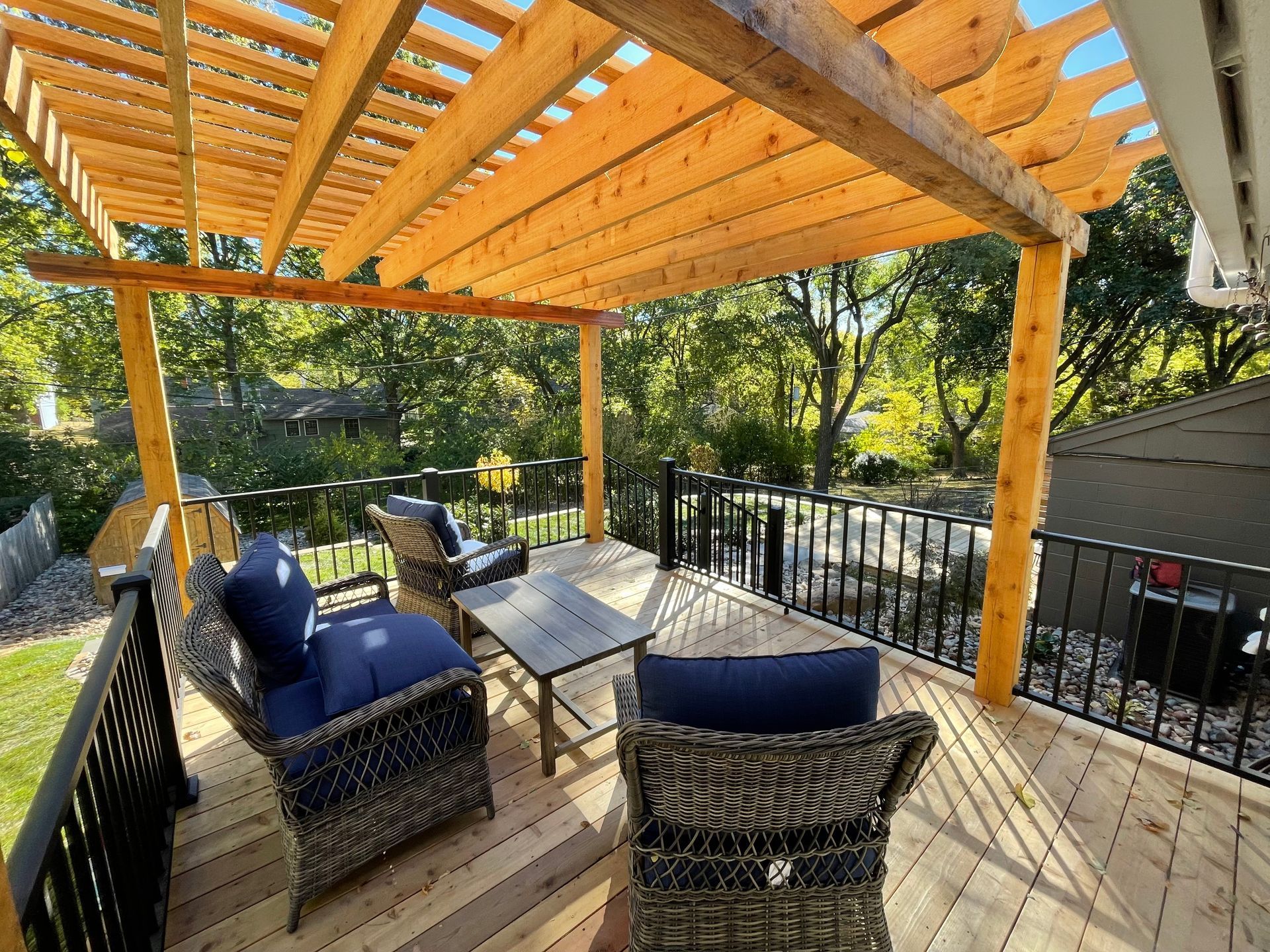 Wooden deck with wicker chairs and a table under a pergola, surrounded by trees and a black railing.