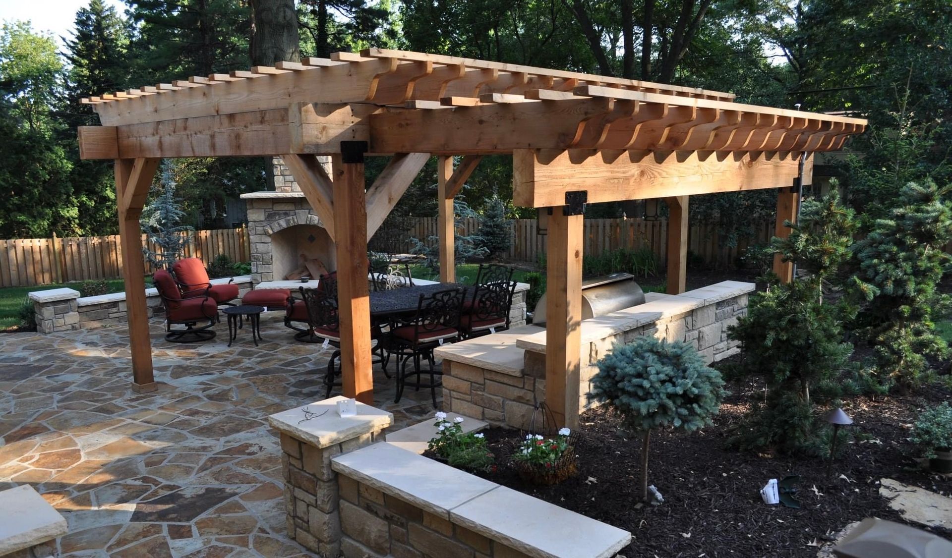 Wooden pergola over a stone patio with outdoor seating and garden beds in a wooded yard