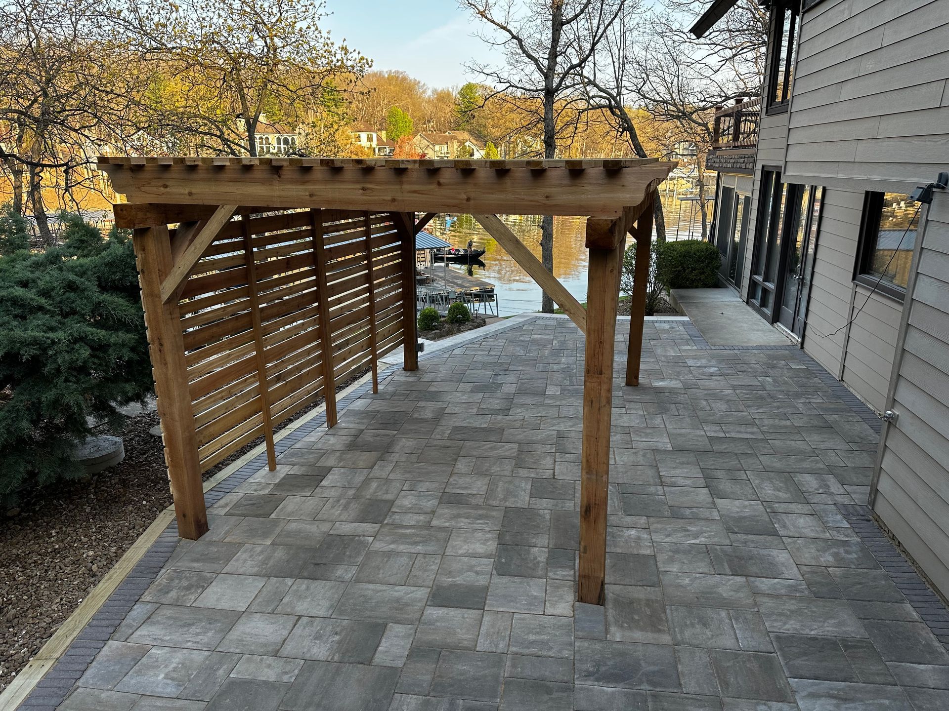 Wooden pergola over a stone patio beside a house, with a sloped driveway and trees in the background