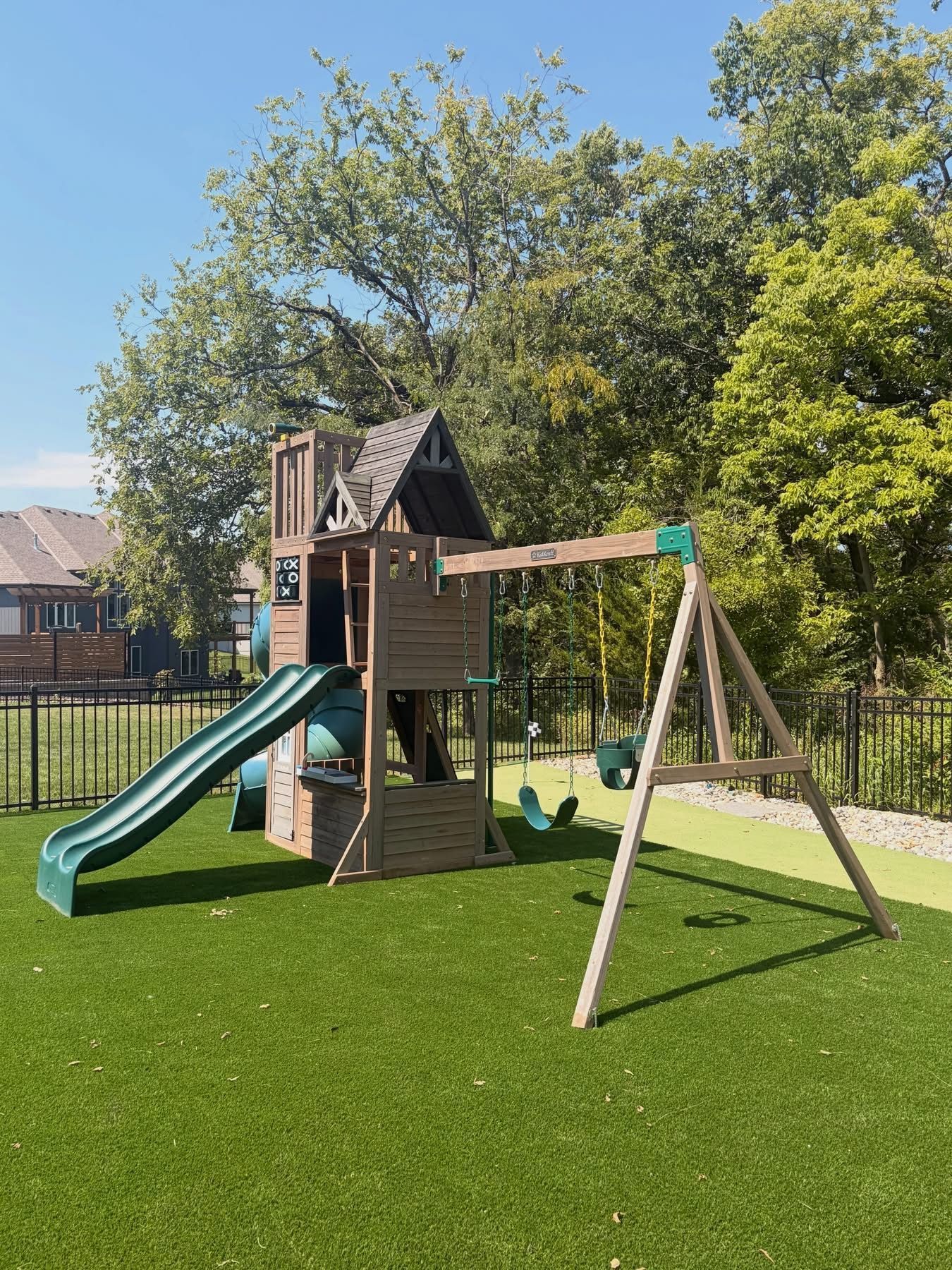 Children’s backyard playset with green slide, tower, swing frame, and large trees on a sunny lawn