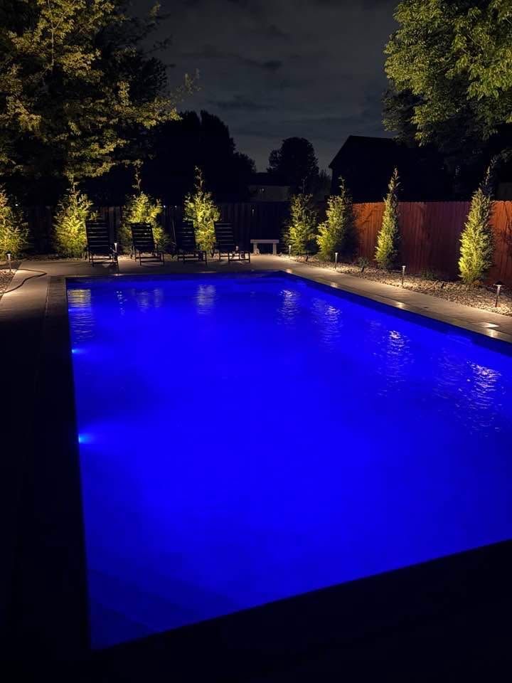Backyard swimming pool glowing blue at night, surrounded by lit trees and a dark sky