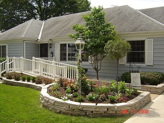 Ramp access to a light blue house, surrounded by a stone-edged garden with flowers. Green lawn and a small tree.