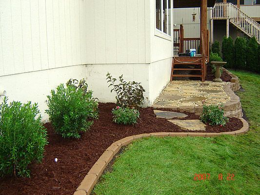 Flower bed with mulch, brick border, and greenery next to a white house with steps leading to a deck.
