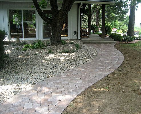 Brick pathway leading to a house with a porch, flanked by gravel and plantings.