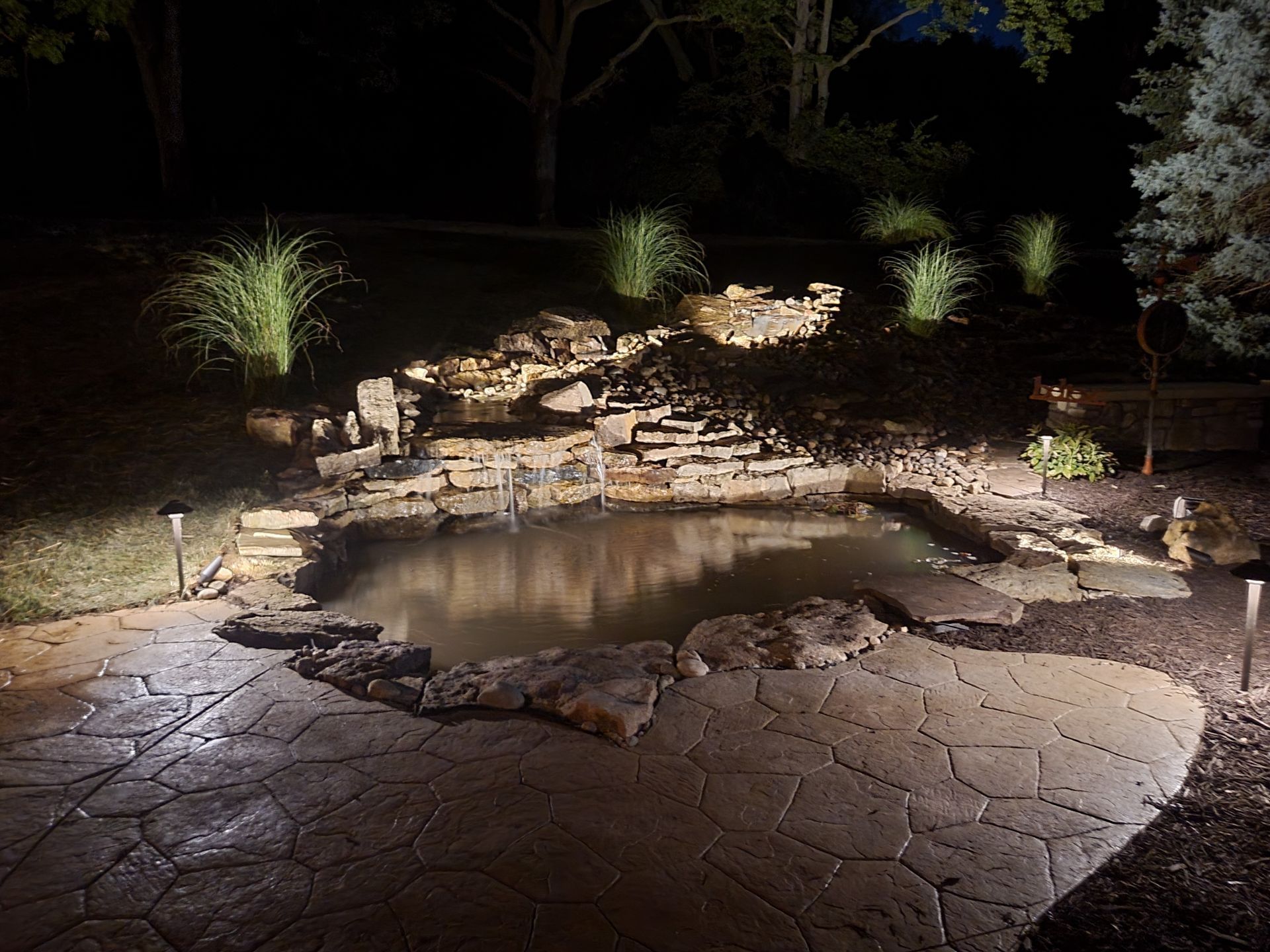 Lit backyard pond with stone border and landscaped rocks at night
