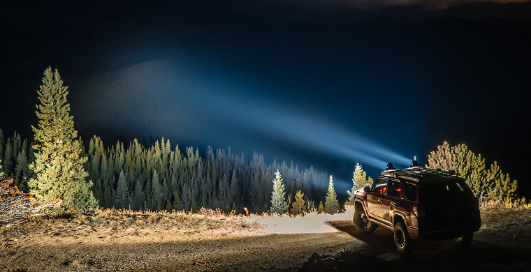 a car is parked on a dirt road at night with a light shining through the trees .