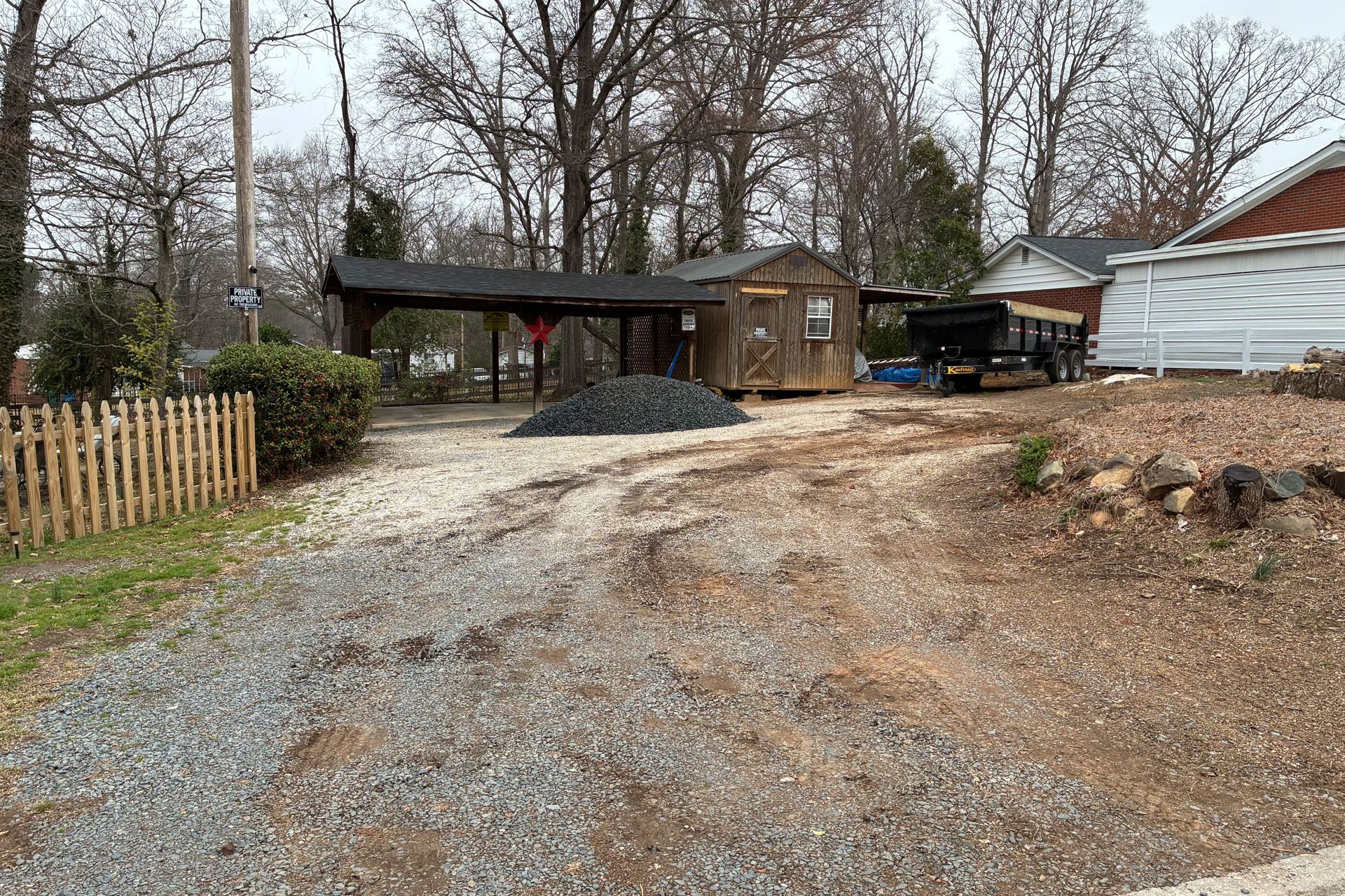 Gravel driveway with a wooden shed and carport. Pile of rocks in the center. Bare trees in the background.