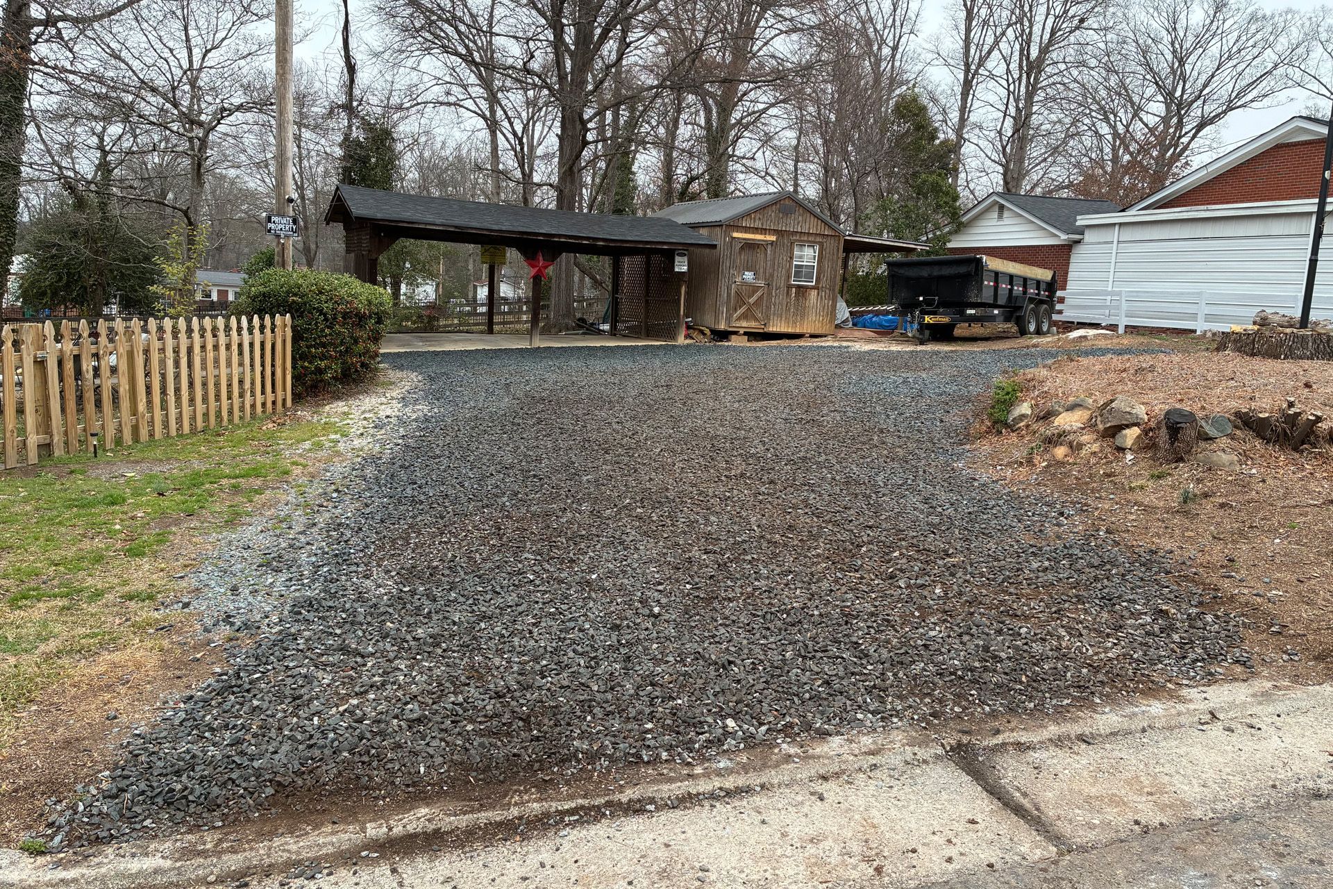 Gravel driveway leading to a shed, carport, and trailer, with a wooden fence to the left.