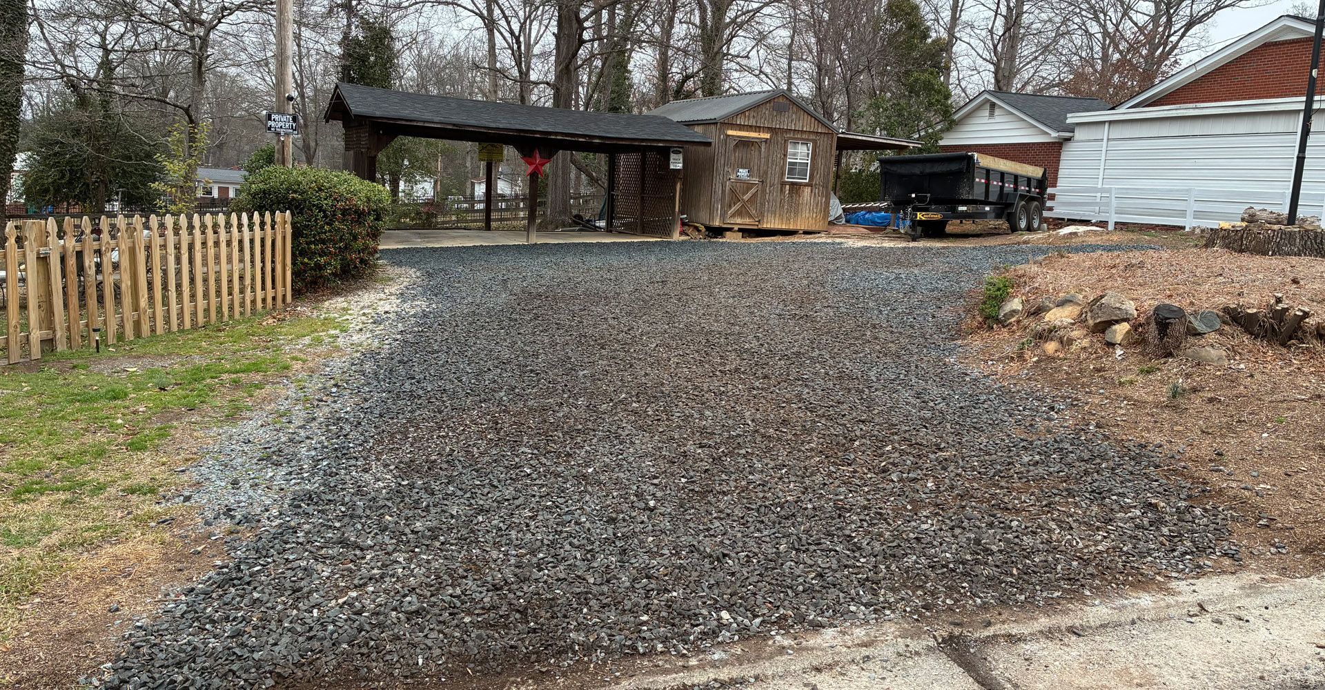 Gravel driveway leading to a wooden shed and covered area. A small wooden fence is on the left.