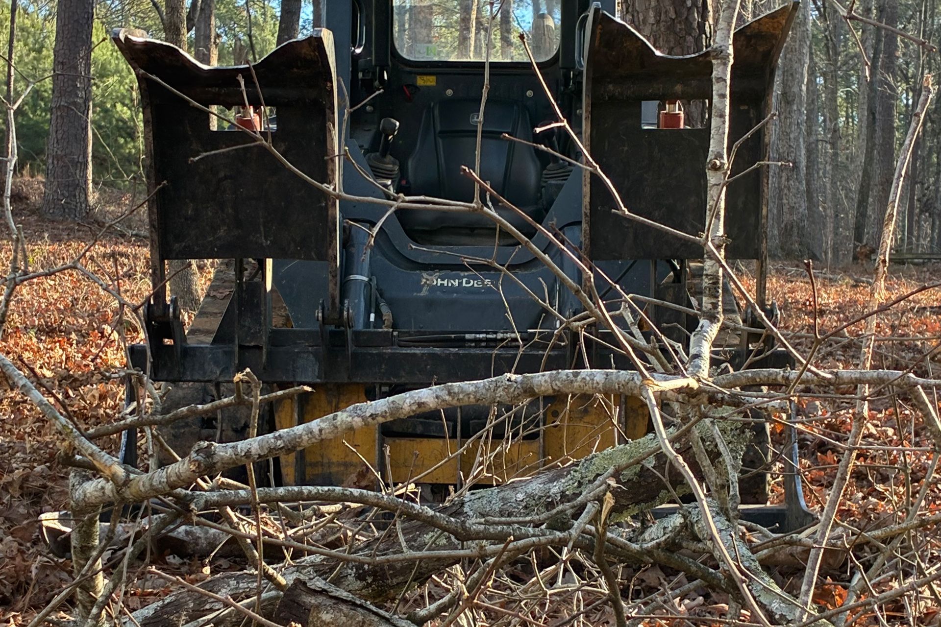 A yellow and black skid steer with a forestry cutter attachment clearing brush in a wooded area.
