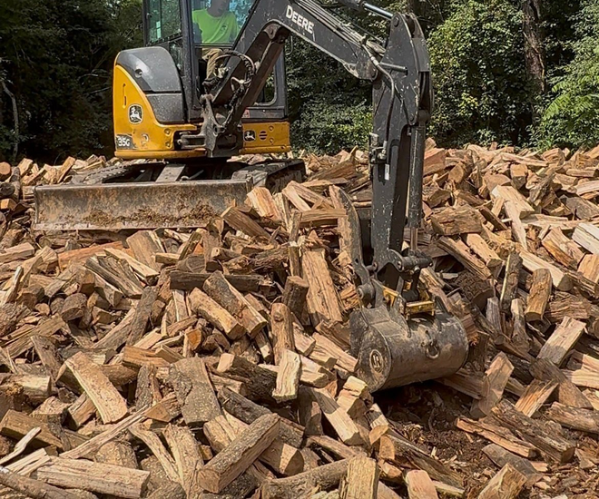 Man splitting wood with a machine outdoors near a John Deere excavator and pile of wood.