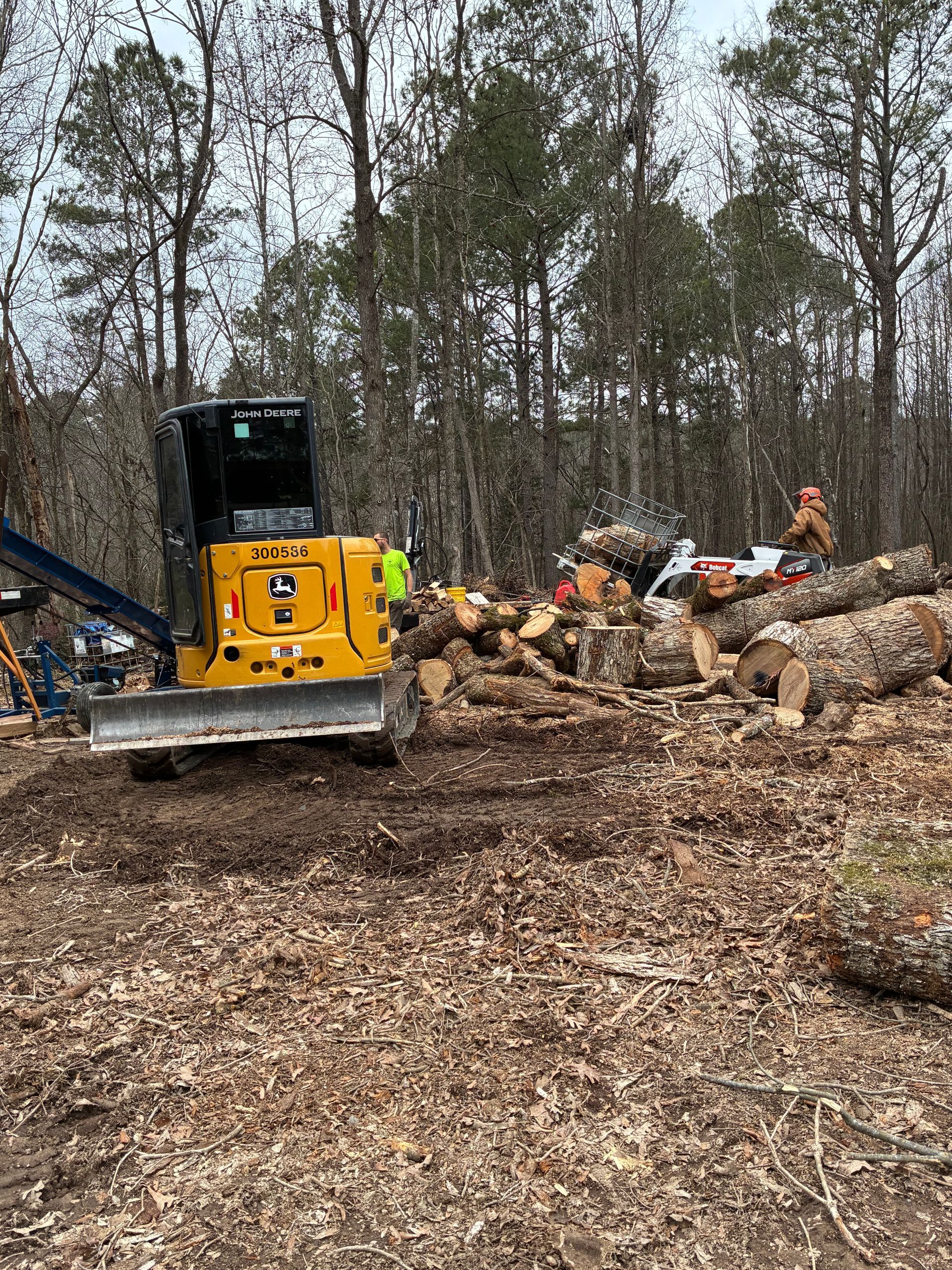 Yellow John Deere bulldozer pushing logs in a wooded area; person with chainsaw nearby.