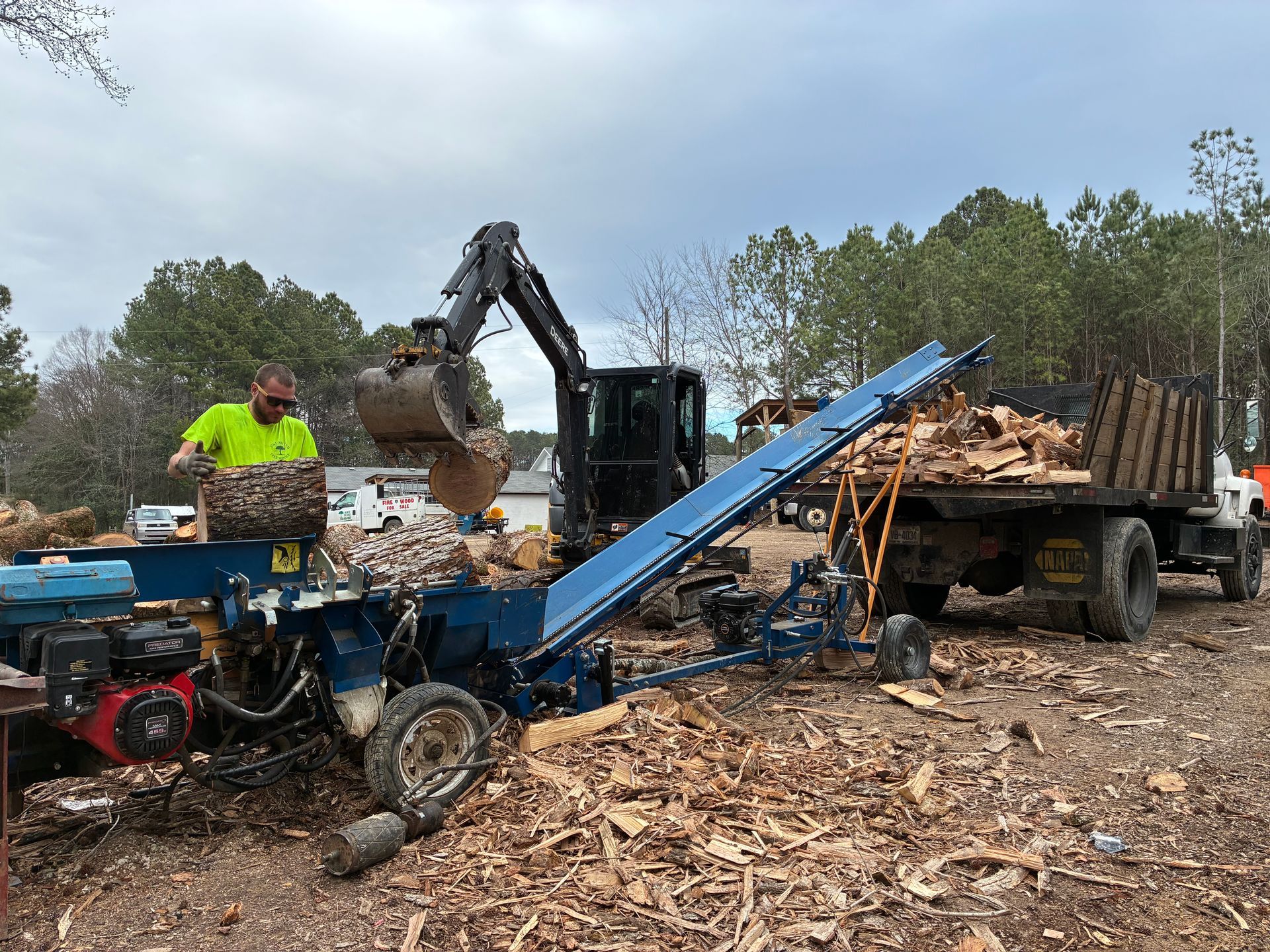 Man loading a wood splitter with an excavator. Logs are split and conveyed into a truck. Outdoors, overcast day.
