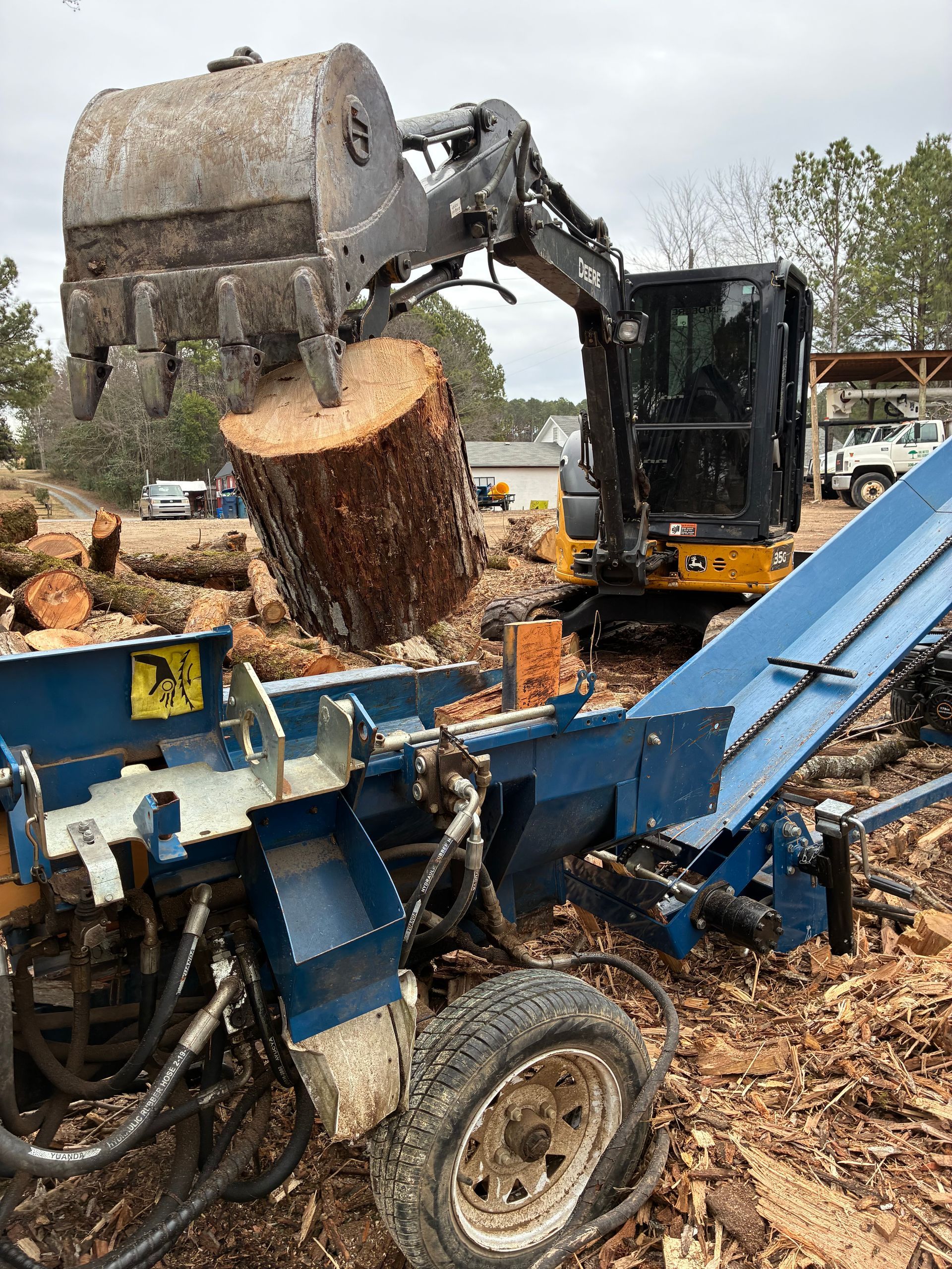Excavator placing a tree log into a blue log splitter. Outdoors.