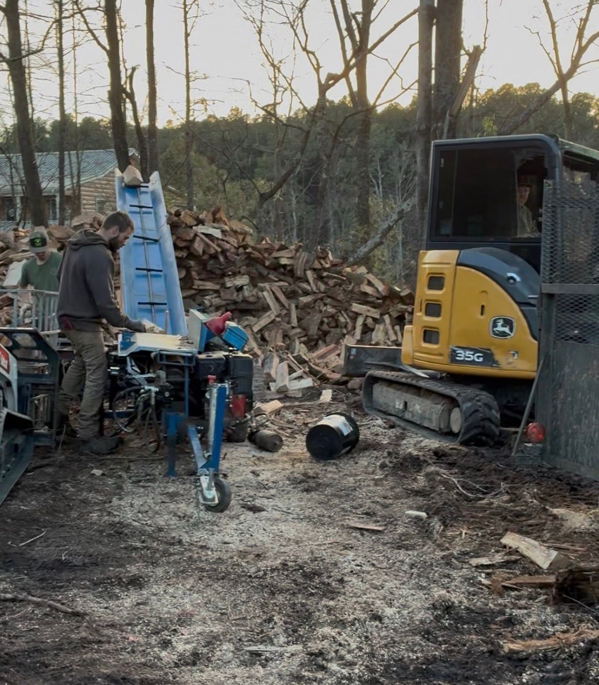 Man splitting wood with a machine outdoors near a John Deere excavator and wood pile.