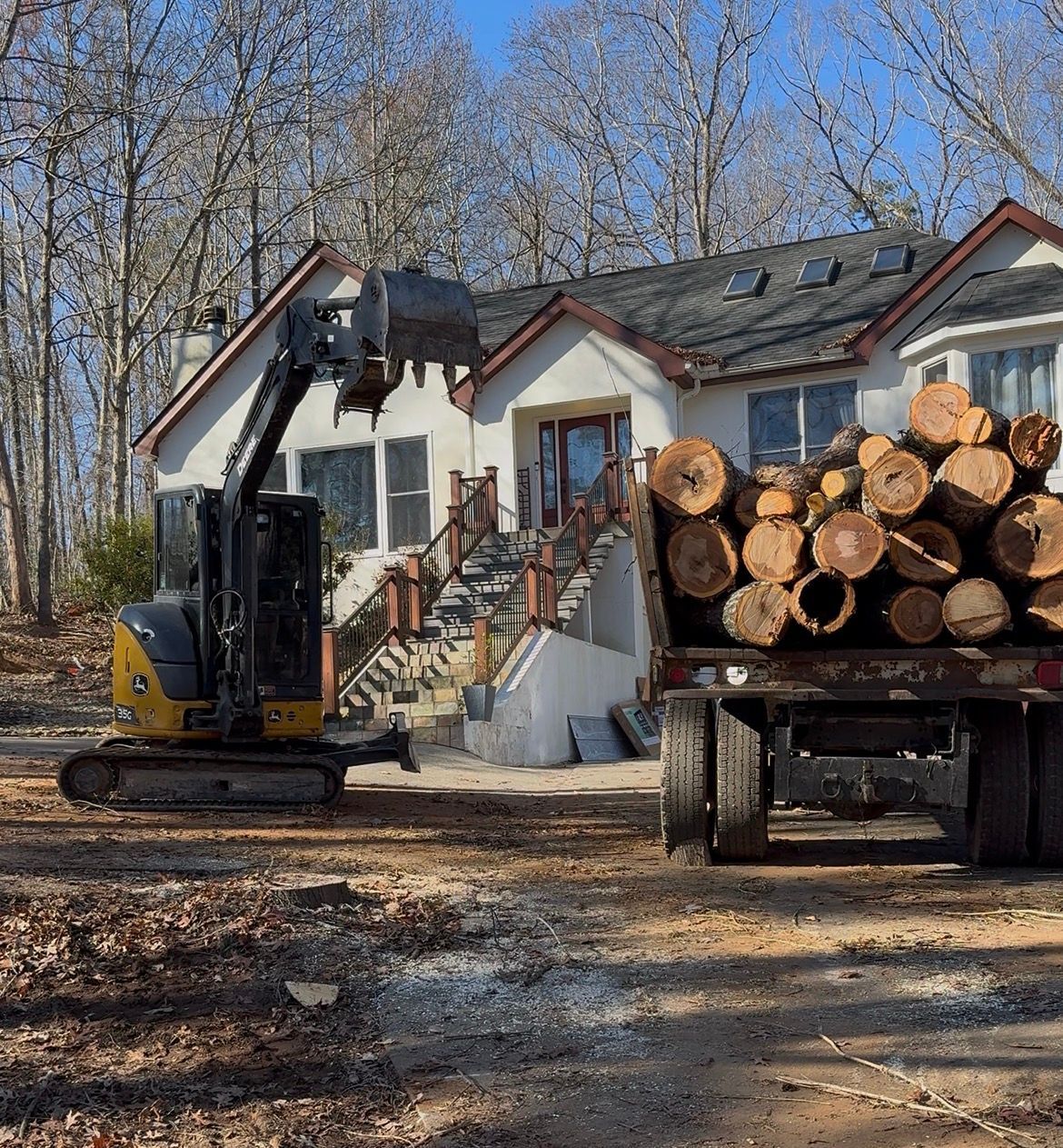 Mini excavator loading logs onto a truck in front of a house.