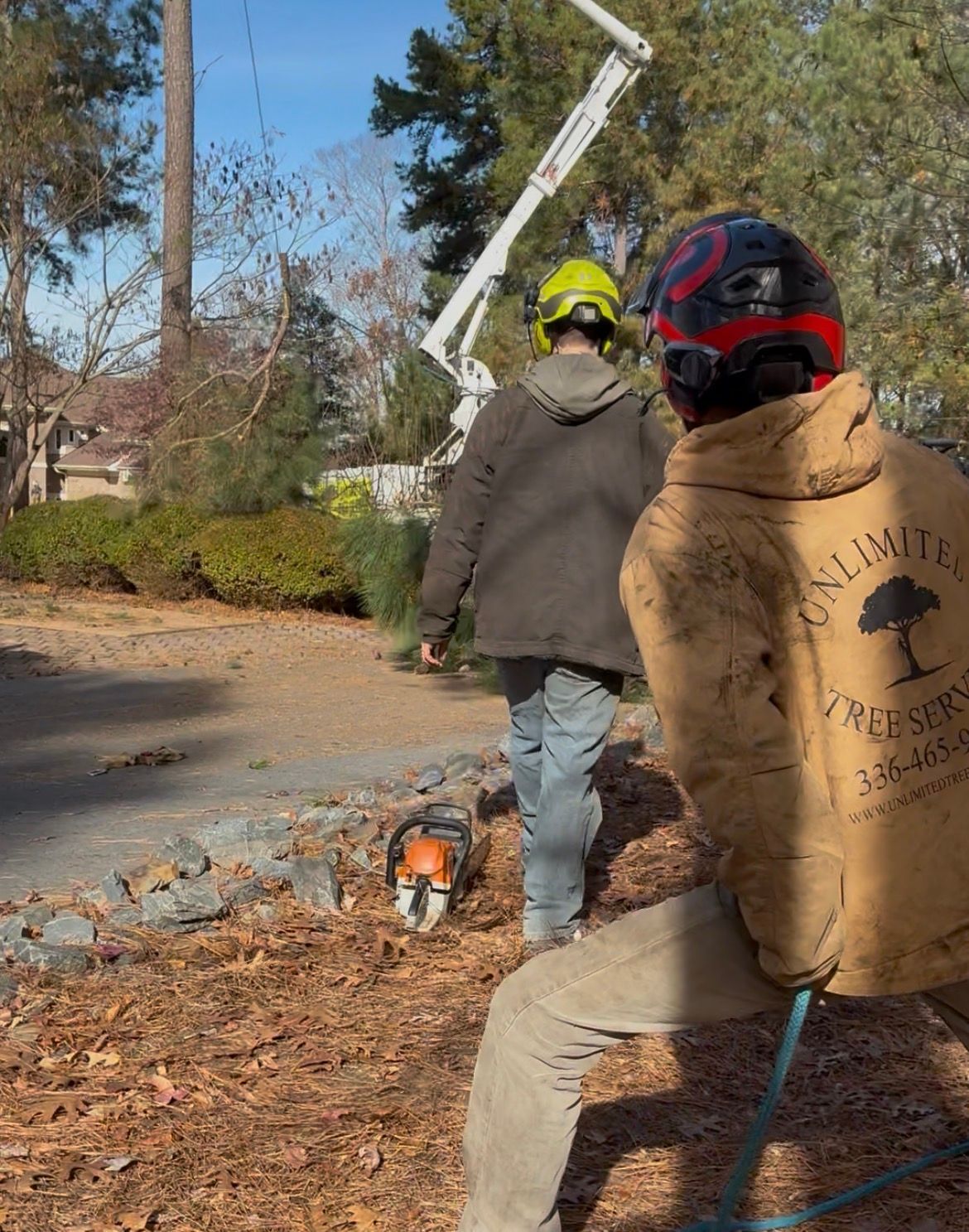 Two tree service workers near a chainsaw. One wears a helmet; the other a company jacket. A boom lift is in the background.