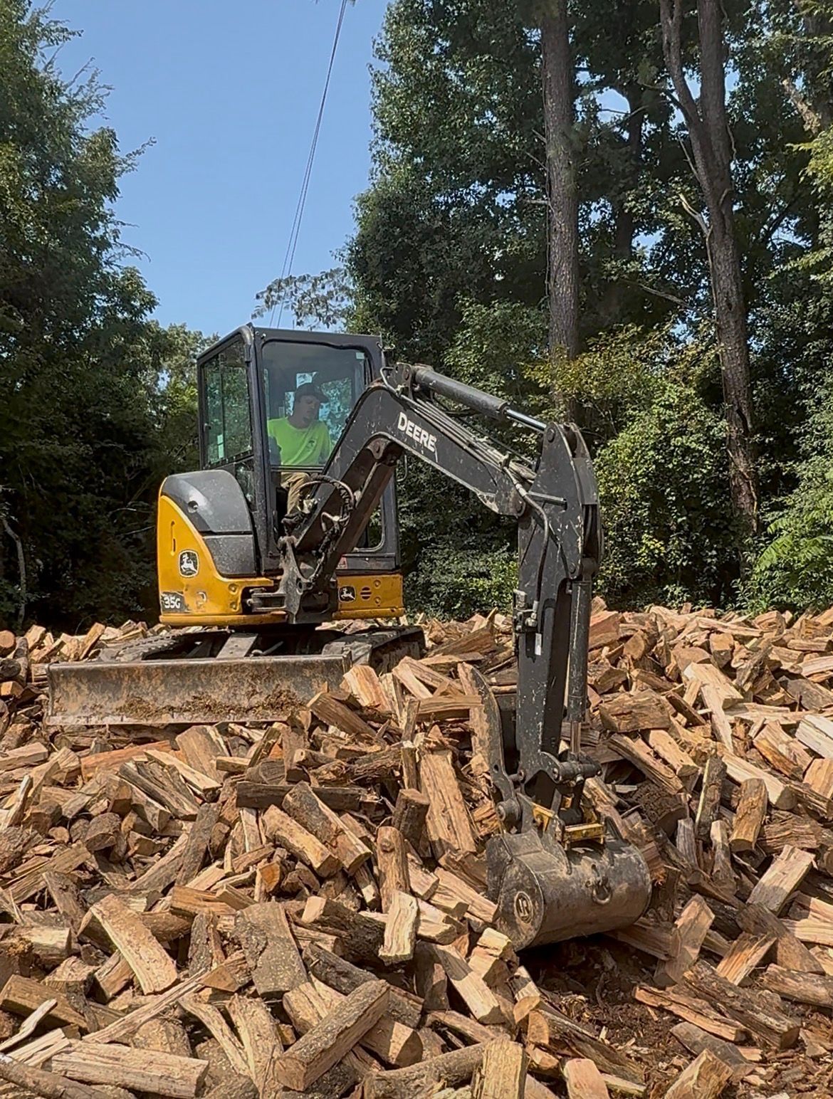 Mini excavator on pile of wood. Yellow and gray machine, green trees in background.