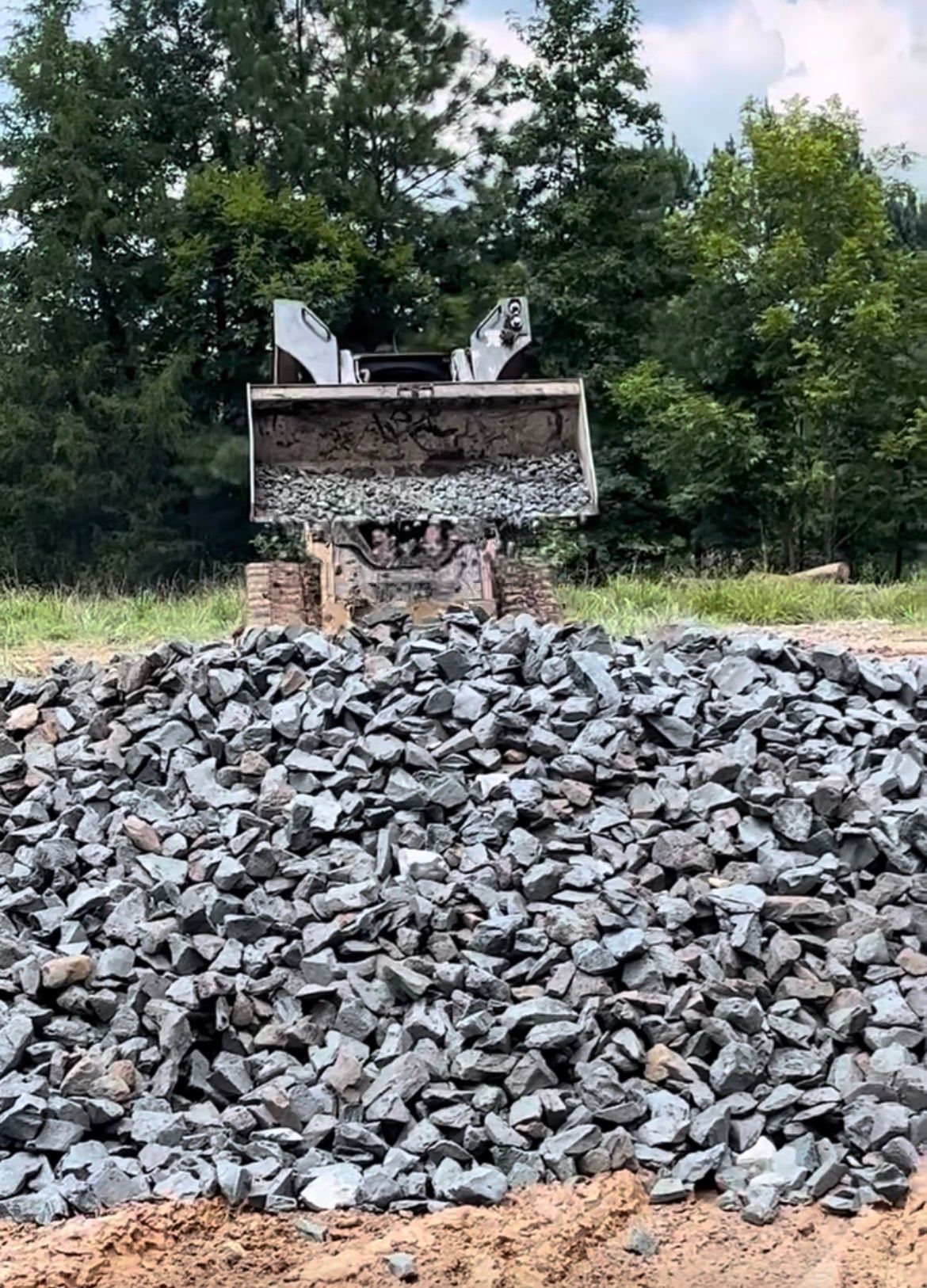 Pile of gray gravel with a front-end loader in a field, trees in the background.