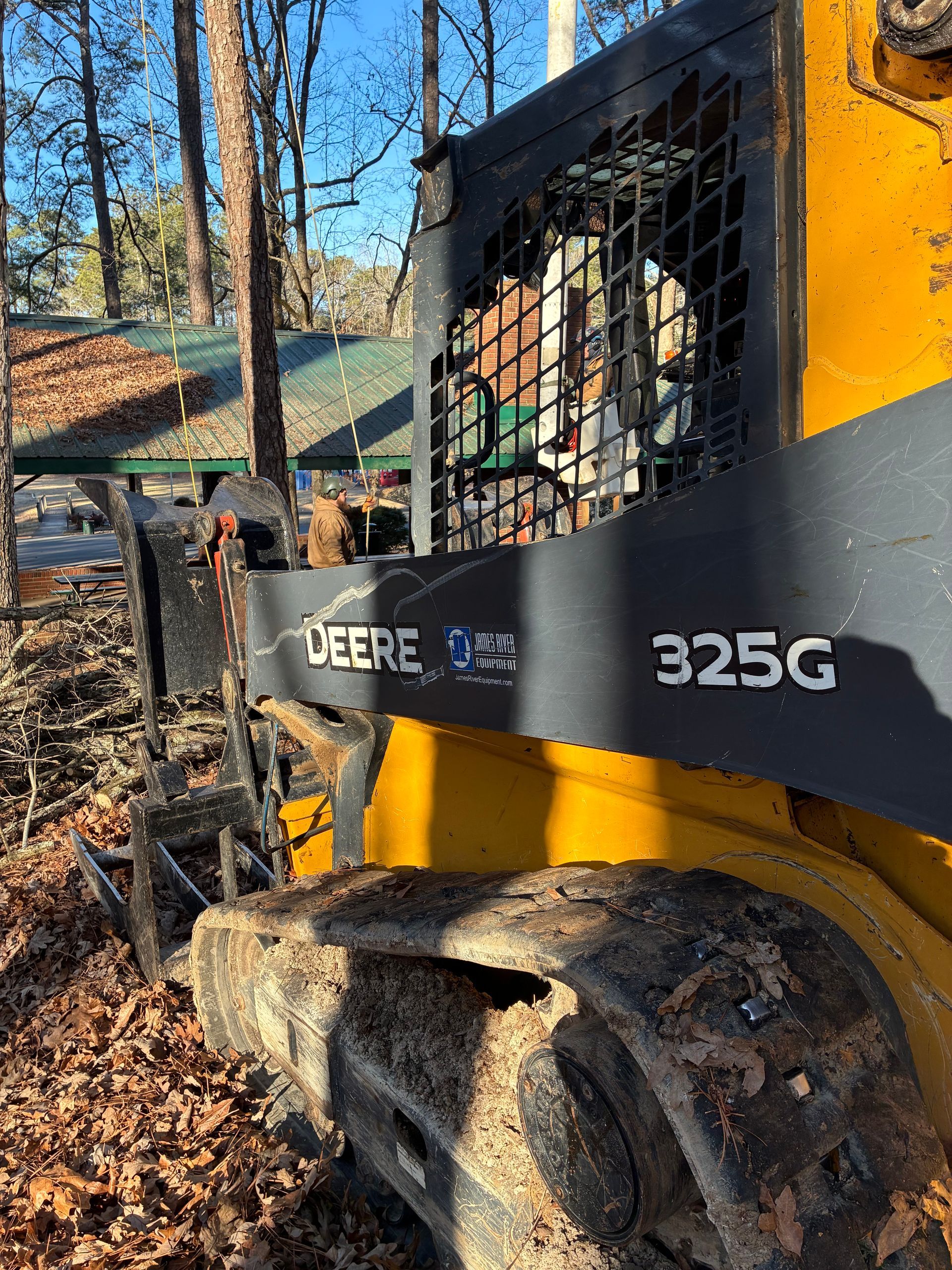 John Deere 325G skid steer loader in a wooded area, operating. Yellow and black machine.