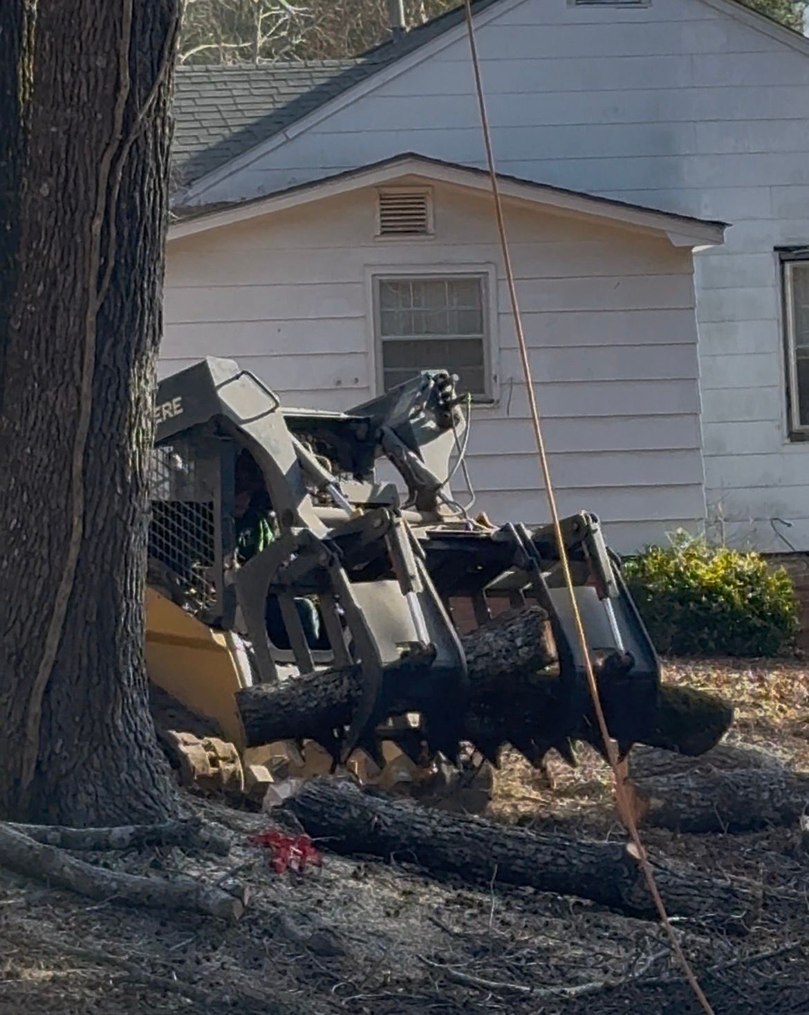 Skid steer with grapple attachment gripping a log near a white house.