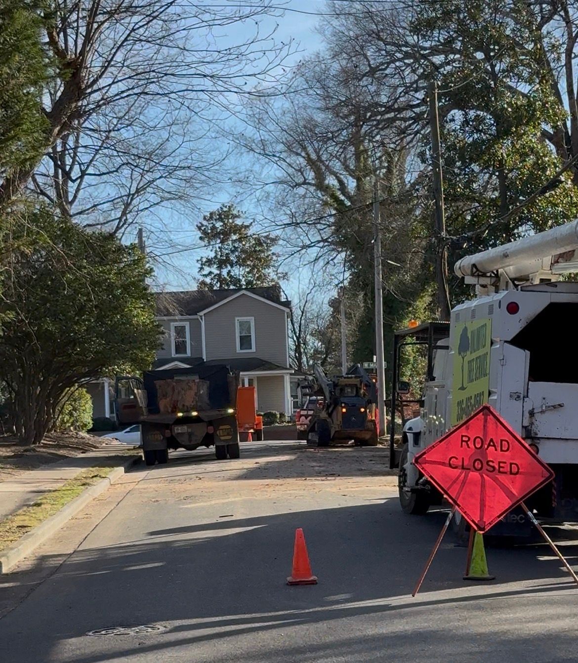 Road closed sign in front of construction. Trucks, equipment, and house visible on a street.