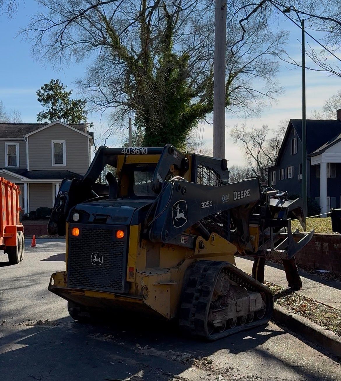 Yellow John Deere skid steer on asphalt road next to a curb. Residential houses and sky in the background.