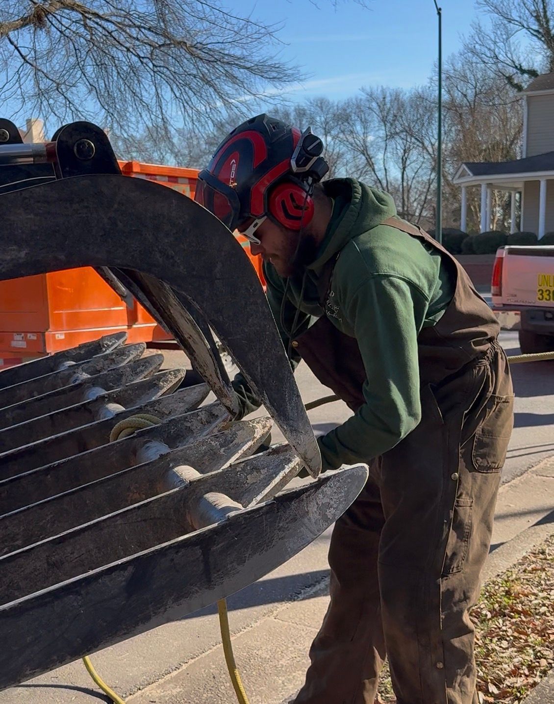 Man wearing safety gear inspects large metal claw attachment on a truck, outdoors on a sunny day.