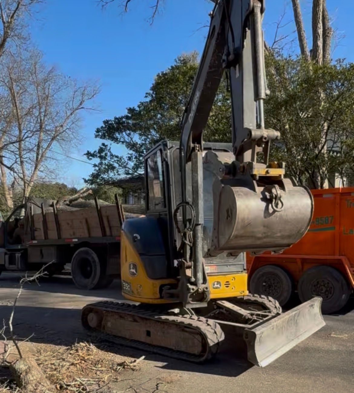 Yellow excavator on asphalt, bucket lowered. A truck loaded with wood debris is in the background.