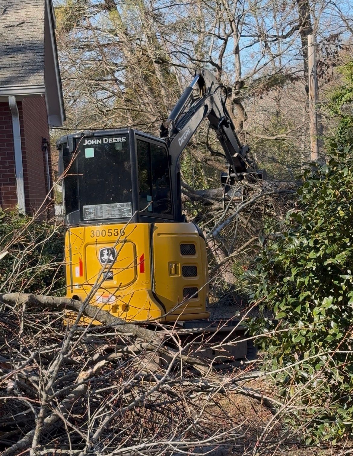Yellow John Deere excavator clearing branches near a brick house.