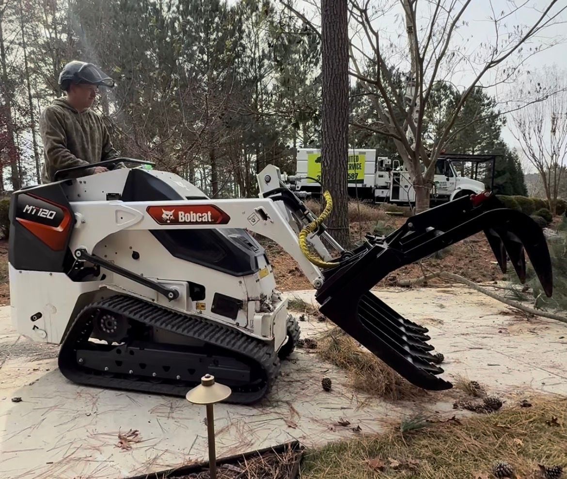 Person operating a white Bobcat tracked skid steer loader with grapple attachment in a wooded area.