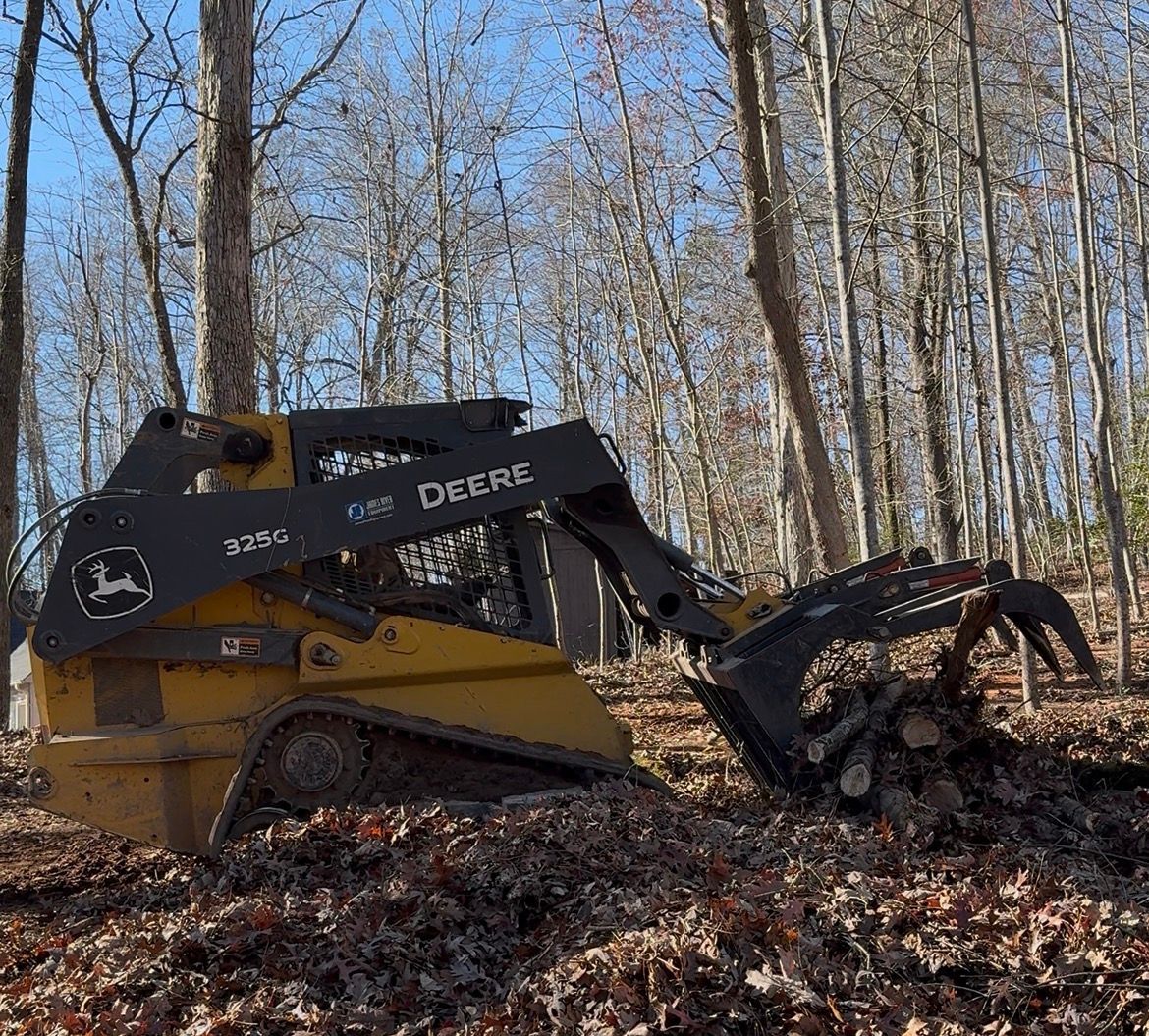 John Deere skid steer with grapple in a wooded area, gathering brush.