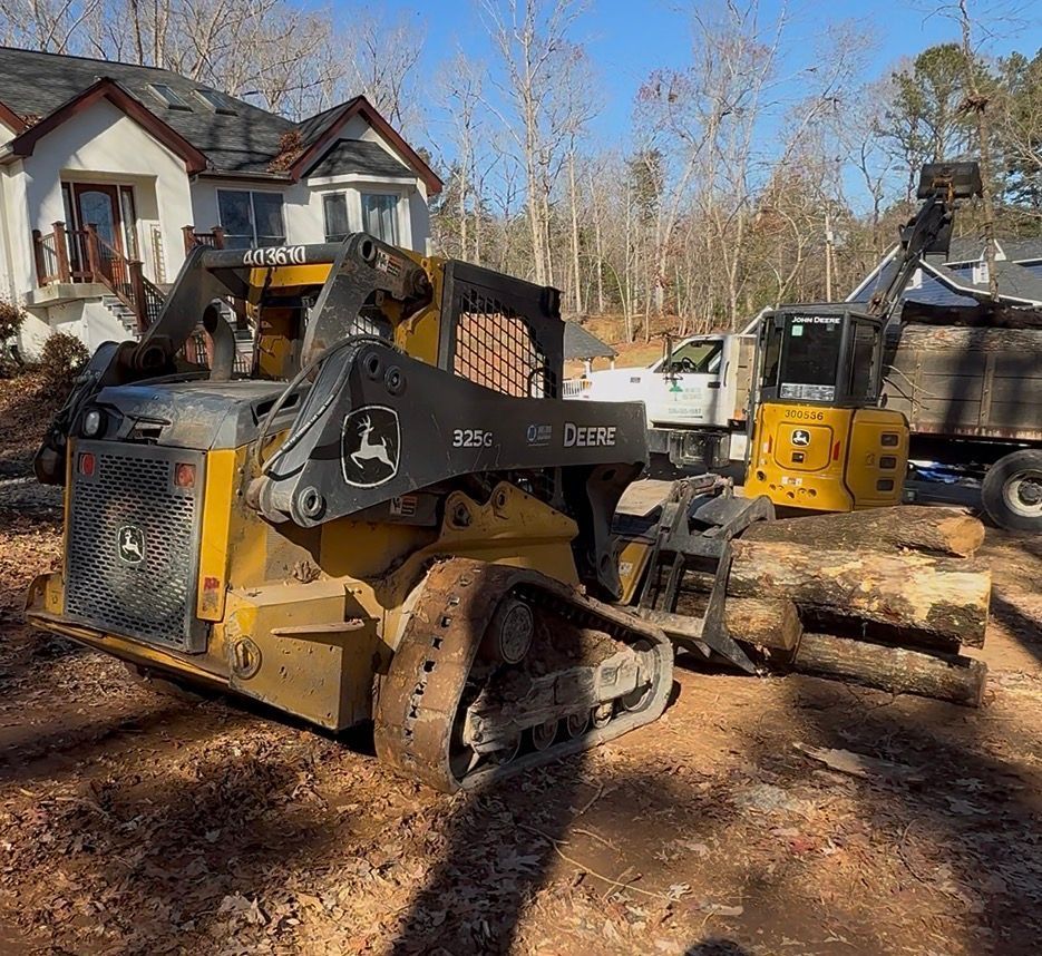 John Deere skid steer and loader moving logs near a house and truck on a sunny day.