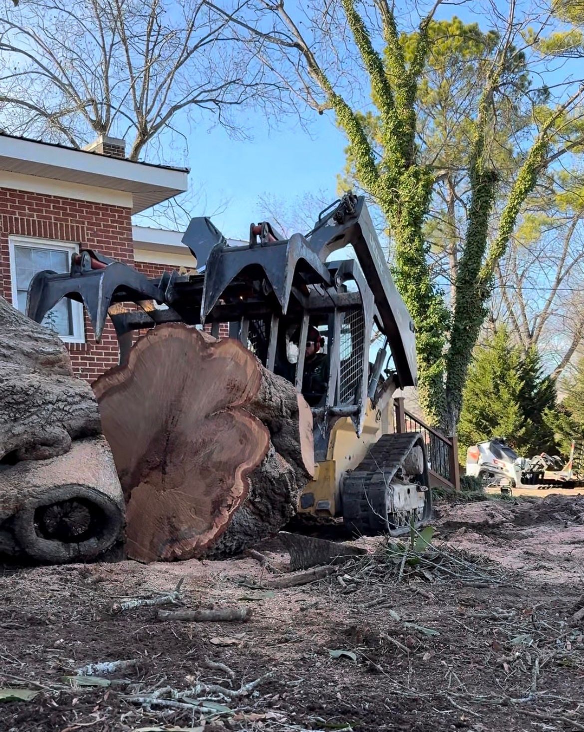 Skid steer with grapple arm lifting a large tree trunk near a brick building.
