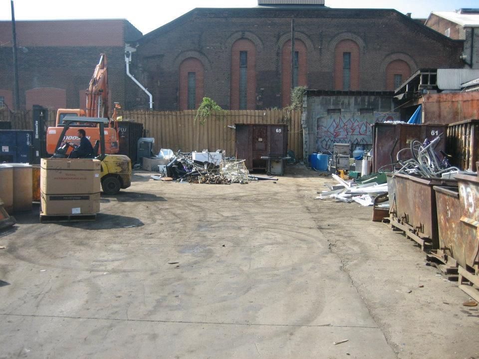 Industrial yard with scattered trash bins and debris in front of a brick warehouse.