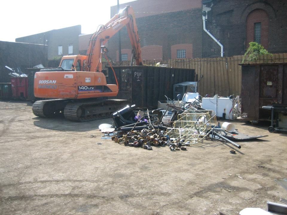 Orange excavator beside a pile of scrap metal in a yard with brick buildings and a wooden fence.