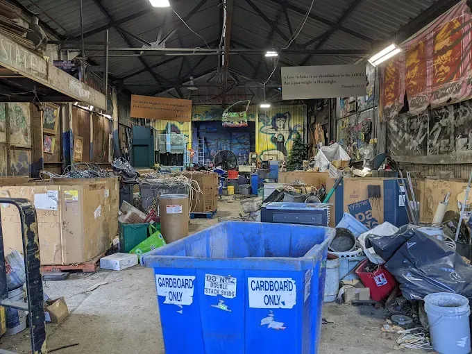 Cluttered warehouse with blue recycling bins and stacked boxes under a metal roof.