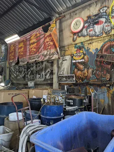 Cluttered workshop with blue bins, hoses, tools, and colorful graffiti-covered walls under a corrugated roof