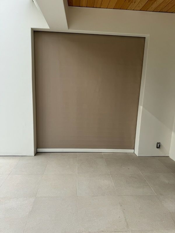 Beige roller shade in an outdoor space, covering a doorway. Light tan tiled floor and white walls.