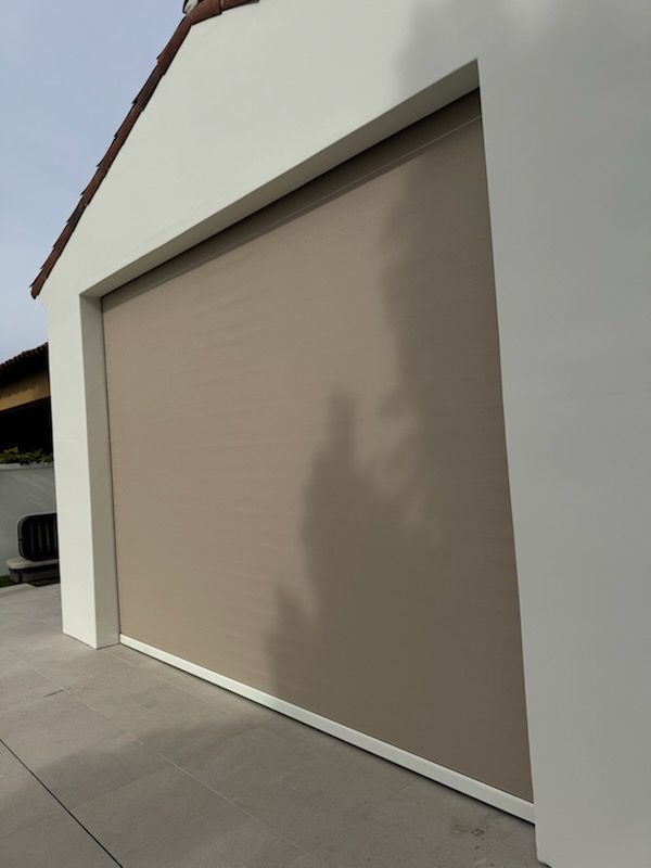 Beige roll-down shade on a white building, casting a shadow. Concrete driveway in front.
