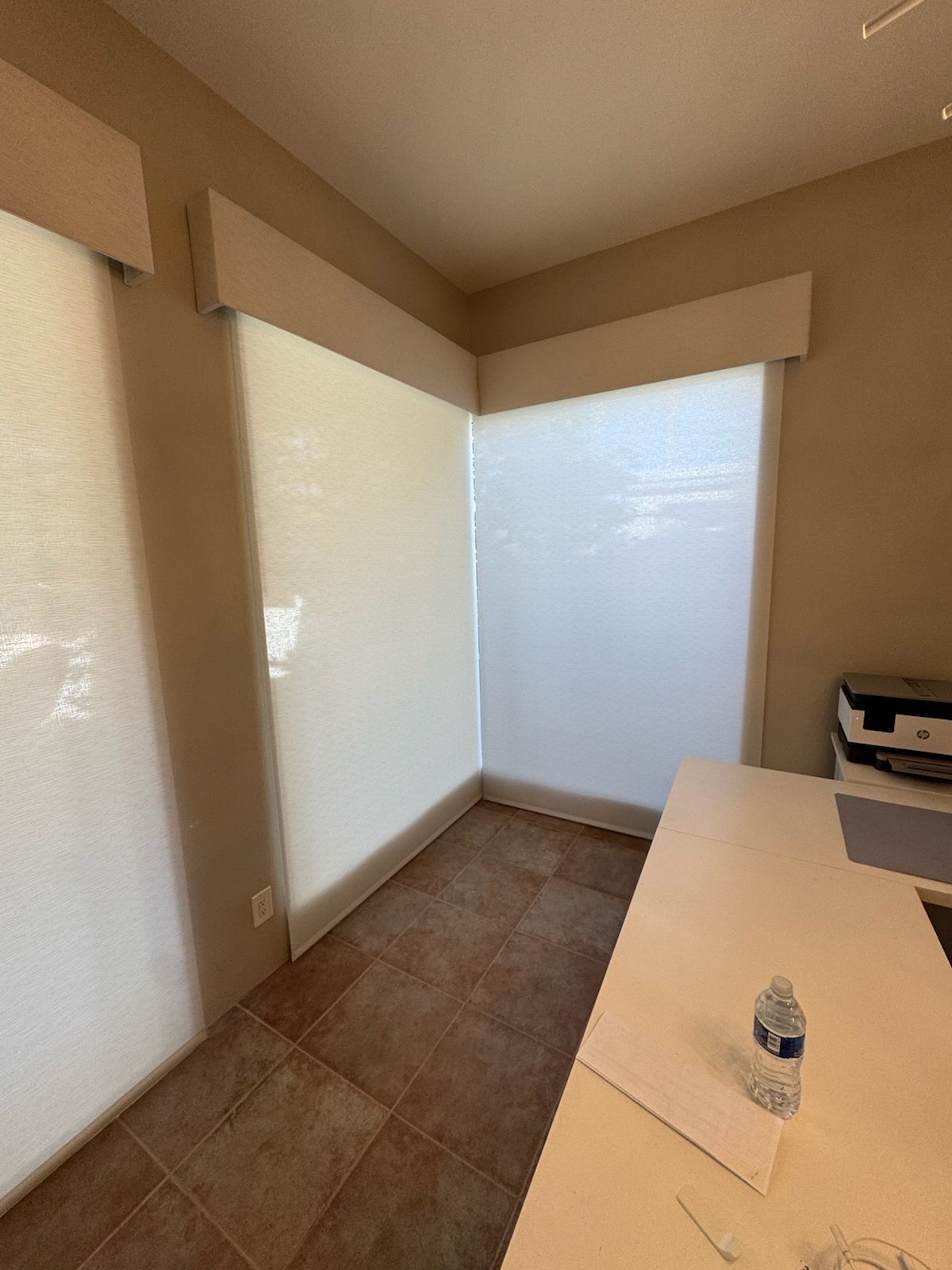 Office corner with white blinds, beige walls, and a white desk. Brown tile floor.