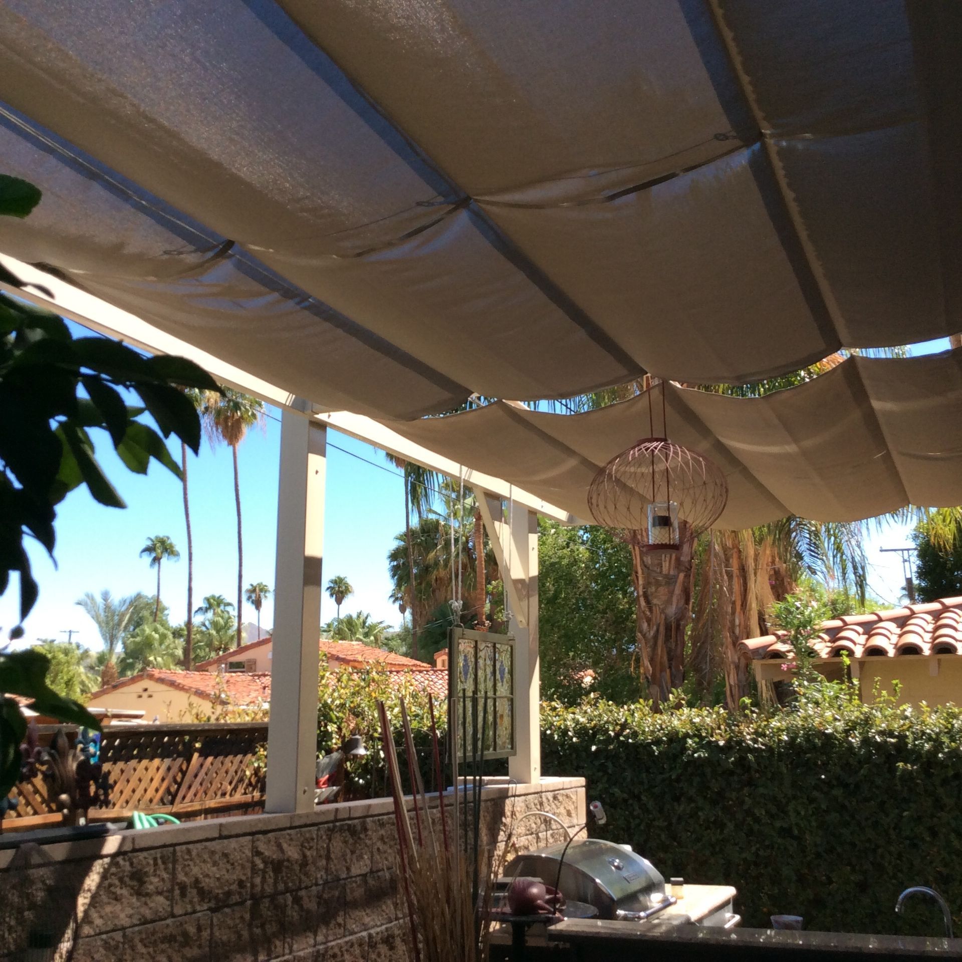 Patio with beige sunshade, grill, and homes with palm trees under a clear blue sky.