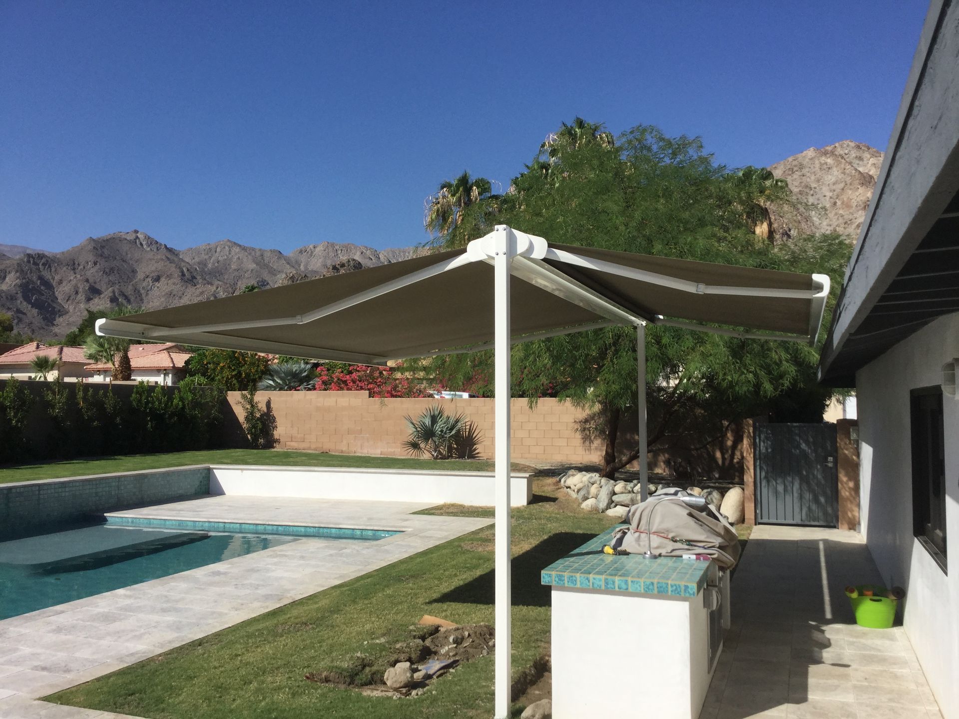 Patio with shade structure over outdoor kitchen and pool, mountains in the background, sunny day.